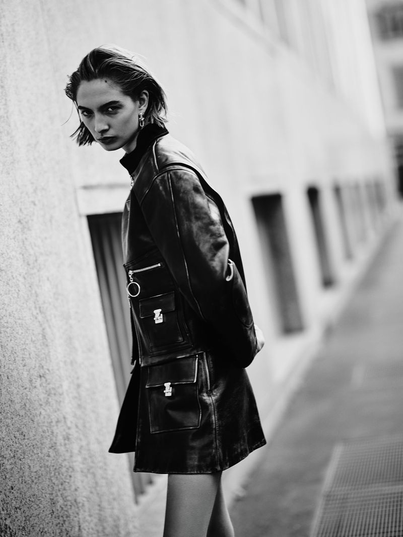 Monochrome portrait of a woman in a black leather outfit standing in front of a building with window openings, styled in an editorial fashion.