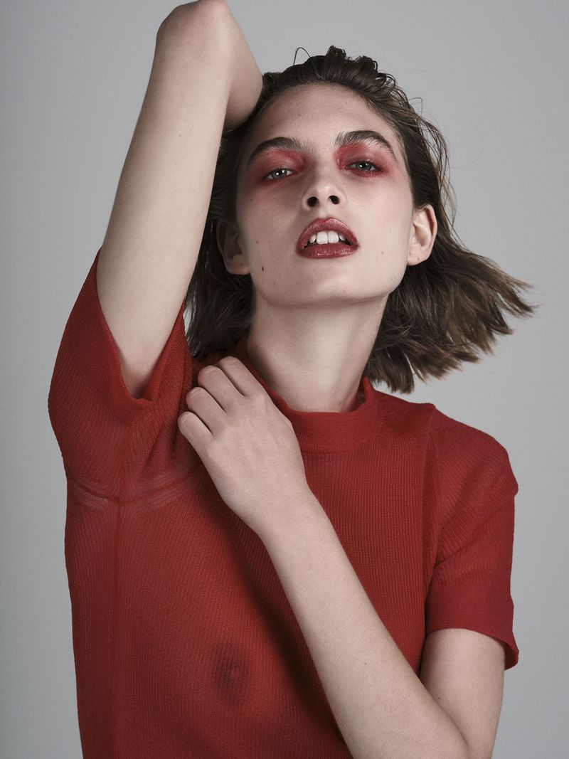 Portrait of a woman with red eye shadow and lipstick in a red shirt against a neutral background, posing with her arm raised.