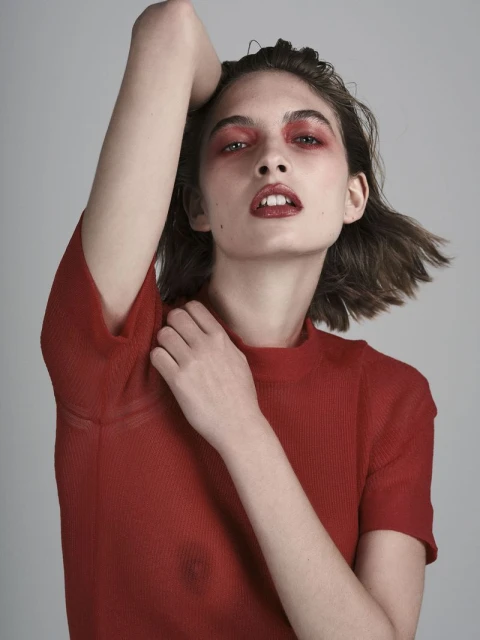 Portrait of a woman with red eye shadow and lipstick in a red shirt against a neutral background, posing with her arm raised.