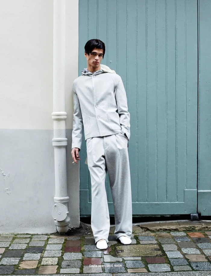 Model wearing a light grey hooded jacket and matching trousers posing against a blue door and pipe in 10 Magazine