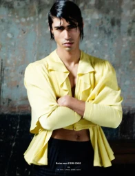 Model with dark wet-look hair wearing a yellow button-up top and black pants against a weathered textured wall background