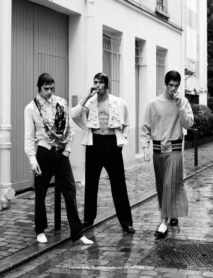 Three male models in contemporary fashion walking and smoking outdoors on a wet cobblestone street lined with buildings.