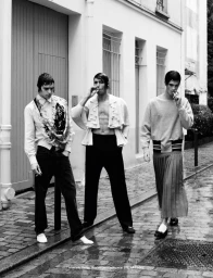 Three male models in contemporary fashion walking and smoking outdoors on a wet cobblestone street lined with buildings.