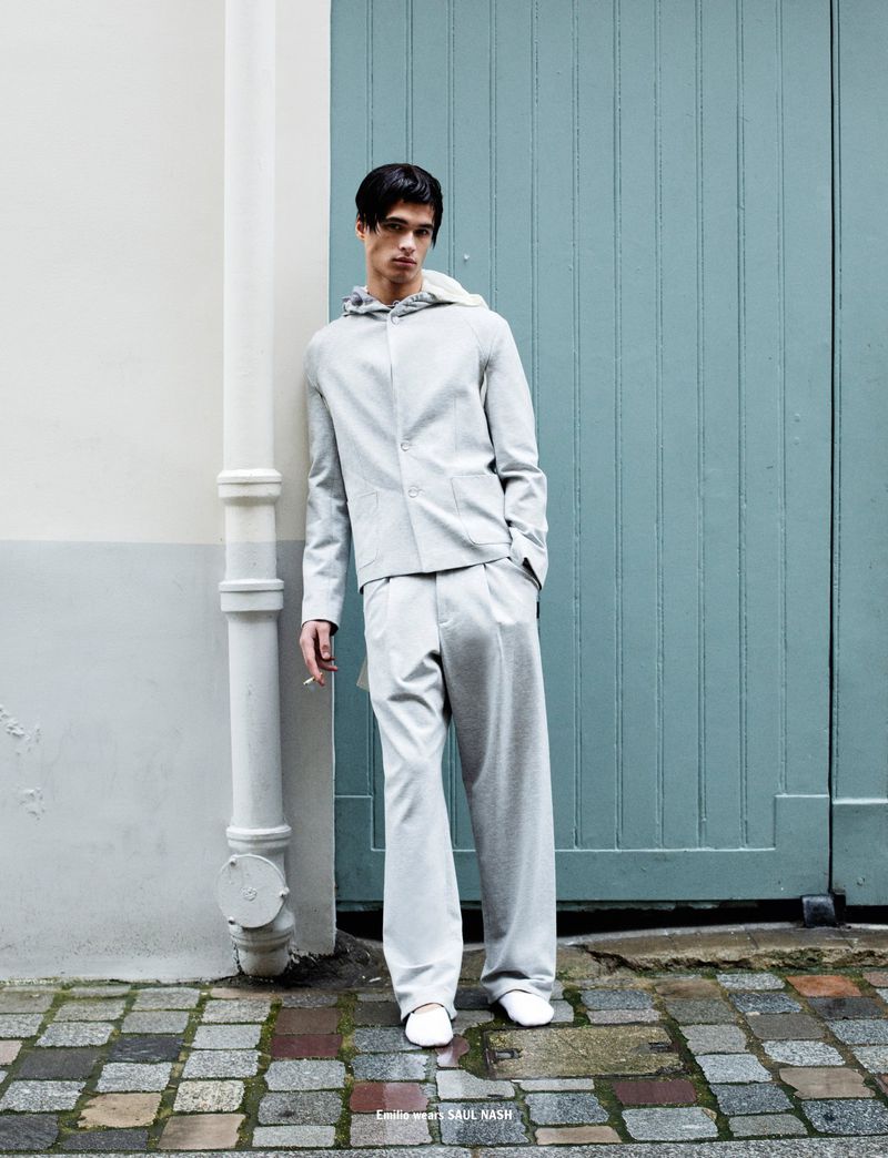 Man poses in light grey hooded jacket and pants, leaning against a blue door on a cobblestone street with cigarette in hand.