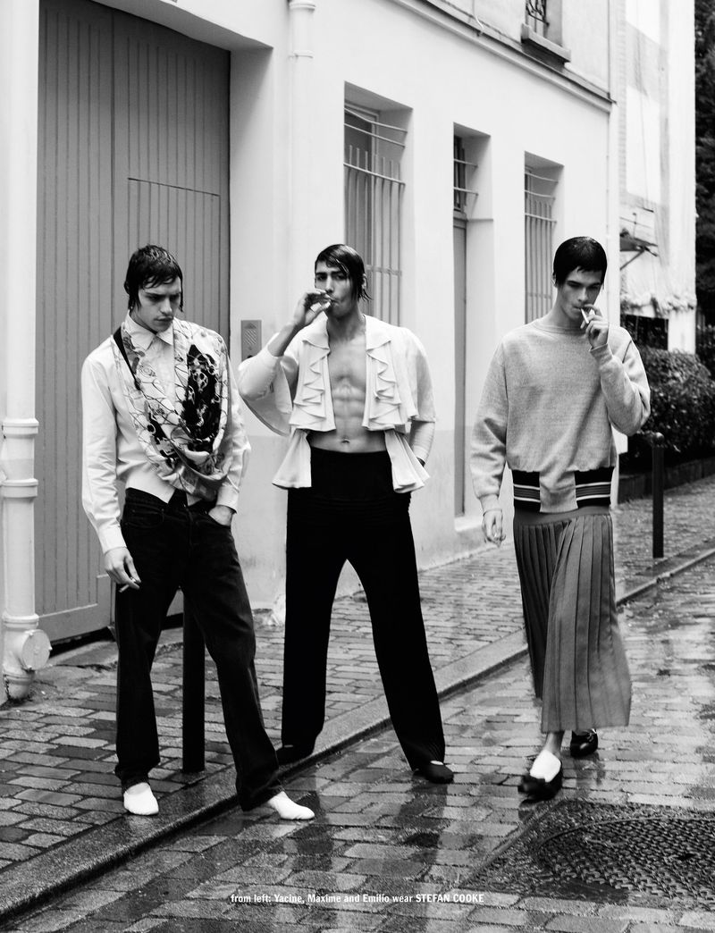 Three adult men pose on a cobblestone street, smoking cigarettes, dressed in stylish clothing, in a black and white fashion editorial.