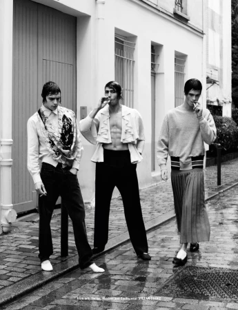 Three adult men pose on a cobblestone street, smoking cigarettes, dressed in stylish clothing, in a black and white fashion editorial.