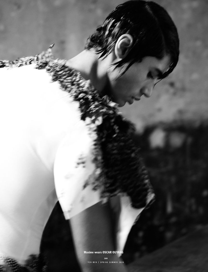 A low angle shot of a stylish male fashion model in a white, modern top with feathered accents, with damp hair, posing indoors.