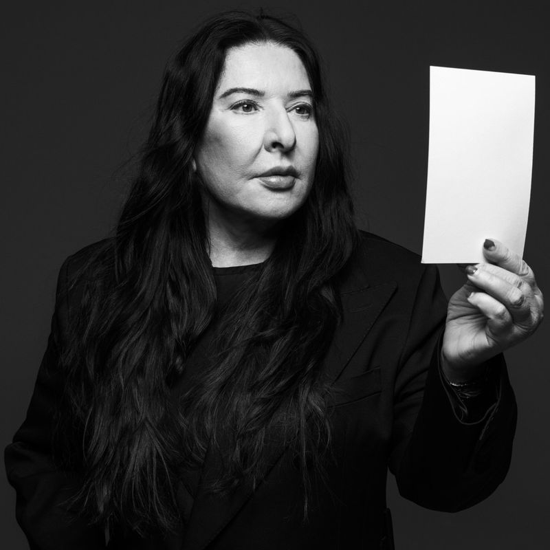 Monochromatic portrait of a woman with long dark hair in a black jacket, holding a white card in a studio setting with a dark background.