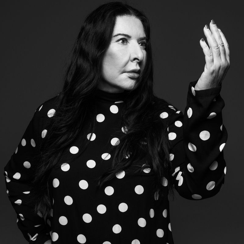 Monochrome portrait of an adult female with long dark hair, wearing a polka-dot dress, raising her hand in a photo studio.