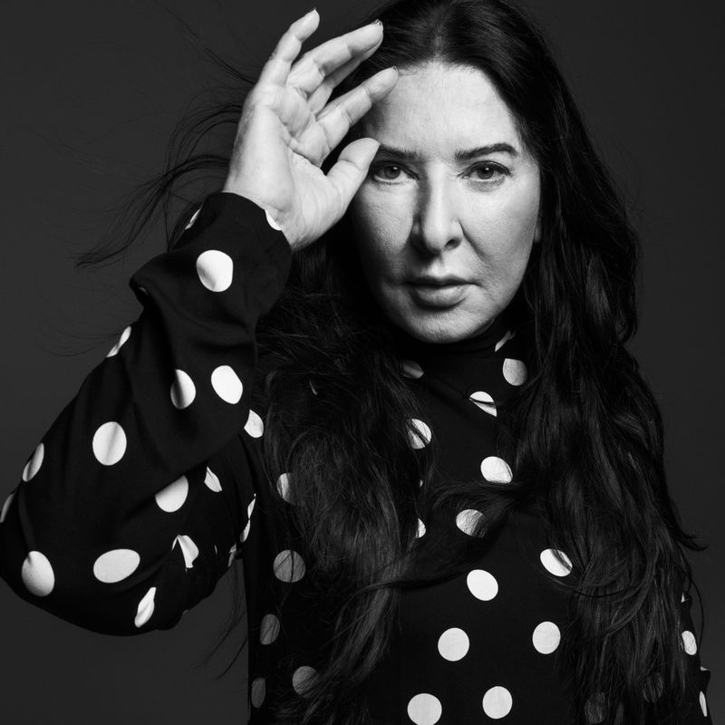 Portrait of a woman with long dark hair wearing a black and white polka dot blouse, looking at the camera with hand near face.