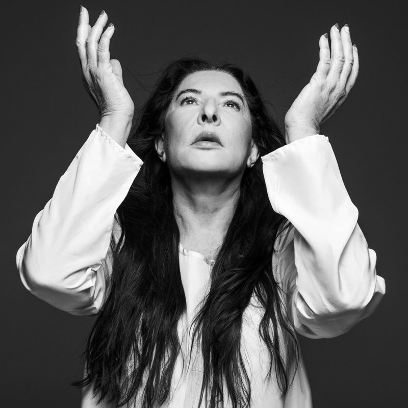 Black and white portrait of a mature woman with long dark hair, wearing a white blouse, with her hands raised, looking up.