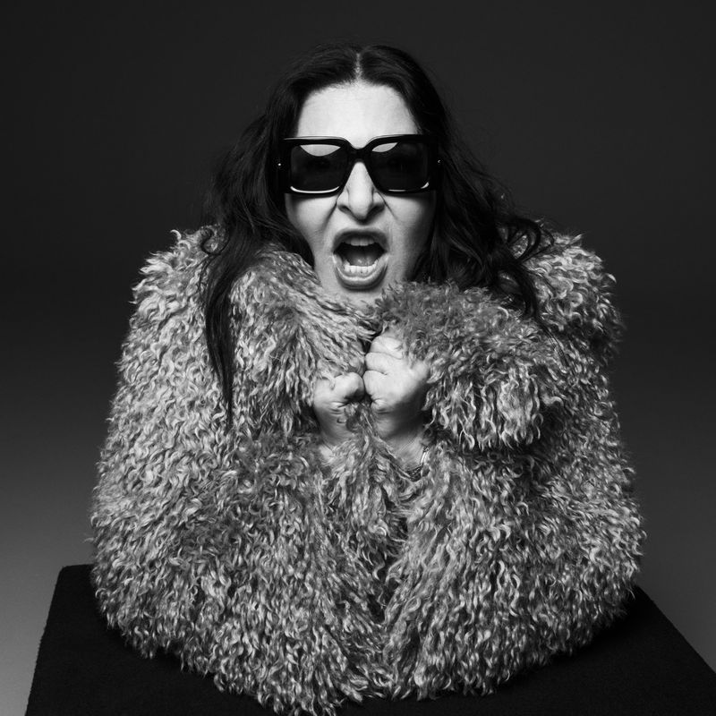 Studio portrait of an adult female with sunglasses and a fur coat shouting with an open mouth in front of a grey gradient backdrop.