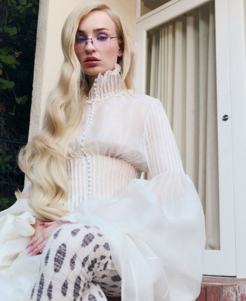 A woman with long blonde hair poses in a sheer white top with a high ruffled collar and glasses, sitting on a porch with green foliage.