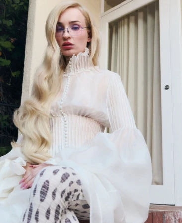 A woman with long blonde hair poses in a sheer white top with a high ruffled collar and glasses, sitting on a porch with green foliage.