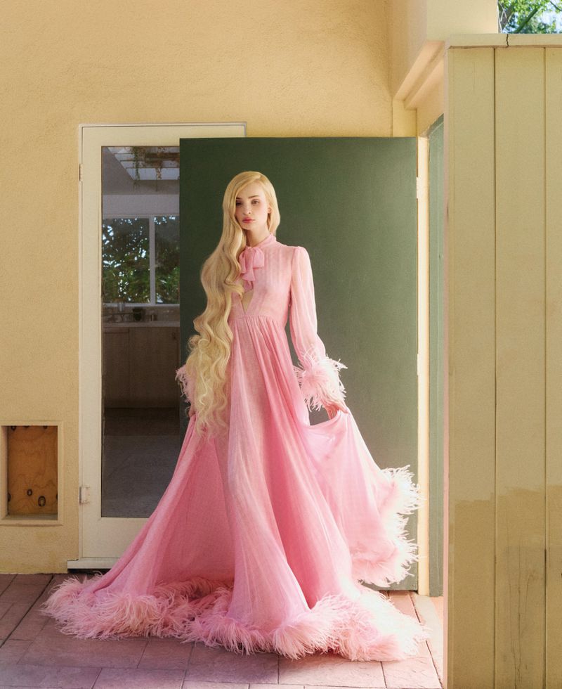 A woman with long blond hair poses in a long pink gown with feather trim, standing in front of a green door and beige exterior wall.