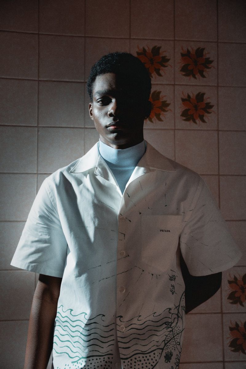 Portrait of Black male fashion model wearing a white Prada shirt with a blue turtleneck, standing indoors against a patterned tile wall.