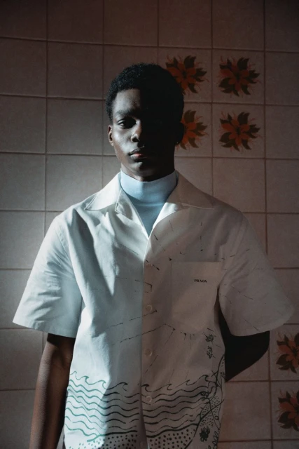Portrait of Black male fashion model wearing a white Prada shirt with a blue turtleneck, standing indoors against a patterned tile wall.
