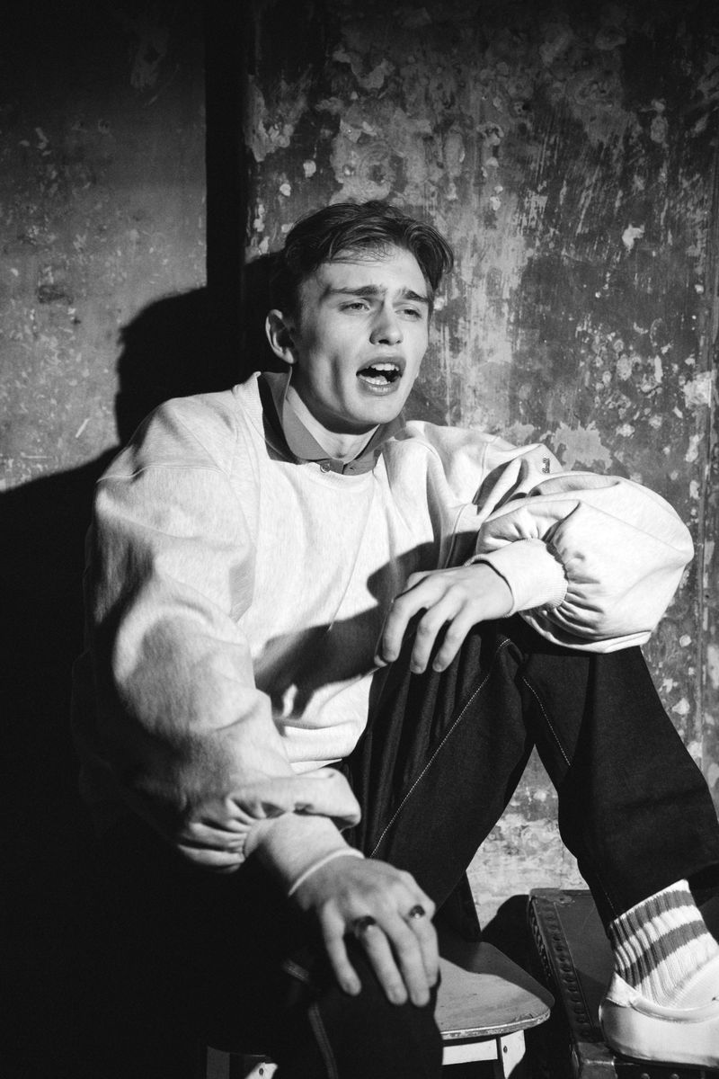 A young man in a white sweatshirt and dark jeans sits on a stool against a textured wall with strong shadows in a black and white portrait.