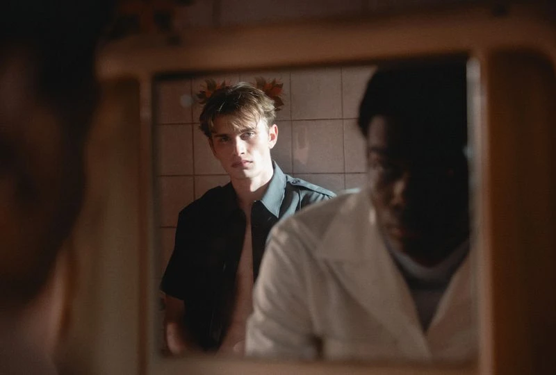Reflection of two young men in mirror, one with dark hair in black shirt, one with dark skin in white shirt, against a tiled wall.
