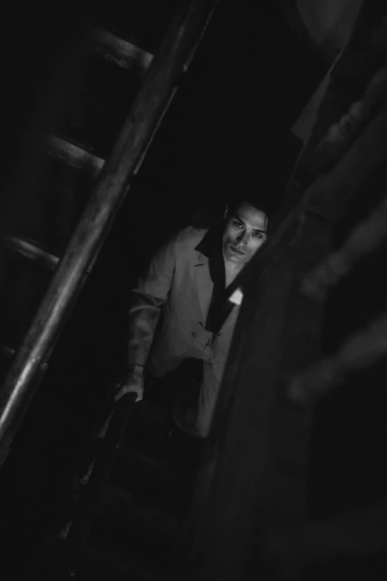Black and white portrait of a man in a suit climbing up a wooden staircase in a dimly lit, indoor environment, captured from a low angle.