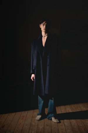 Portrait of a young man wearing a dark overcoat and jeans, standing on a wooden floor with a dark background and dramatic lighting.