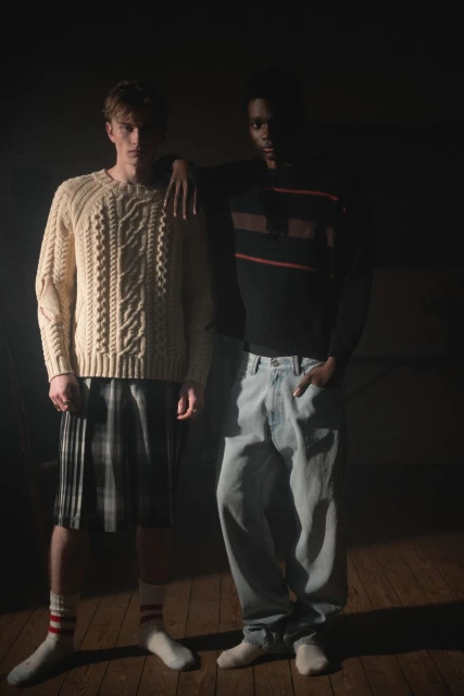 Two men in sweaters and casual pants, one with a cable knit sweater and shorts, and one with a striped sweater and jeans. Indoor shot.
