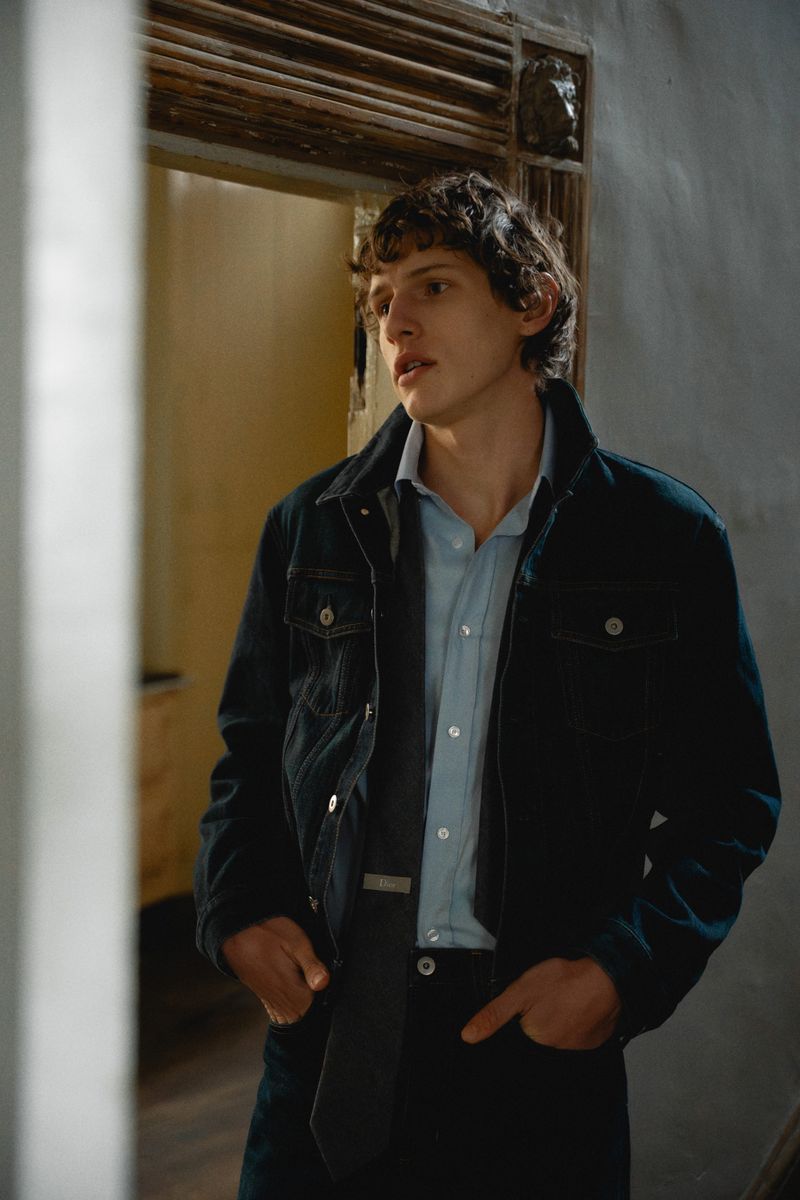 A young man with curly hair wears a dark denim jacket over a light blue button-down shirt, standing with hands in his pockets.