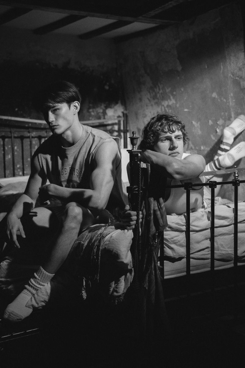 Black and white editorial image of two male models relaxing on an iron bed in a moody room for HERO Magazine.