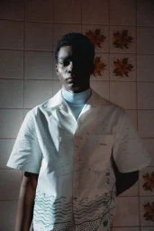 Black man wearing a white patterned short sleeve shirt and light blue turtleneck standing against a tiled wall background