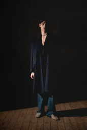 Model standing indoors wearing a dark coat and blue jeans under low-key dramatic lighting on a wooden floor.