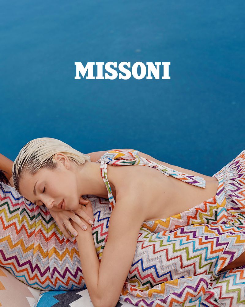 Blonde woman in a patterned dress laying down by a blue background, showcasing a relaxed pose and the 'Missoni' brand logo.