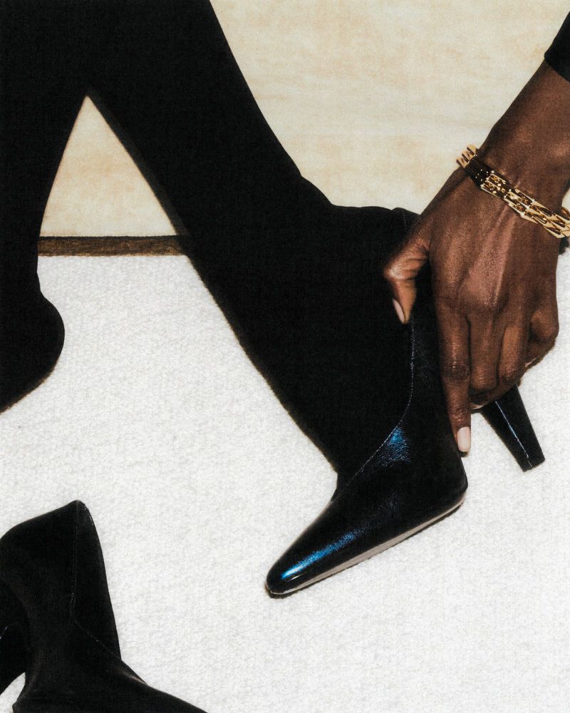 Close-up studio shot of a Black woman wearing a gold bracelet pulling on a shiny black leather ankle boot with black tights on a white carpet.