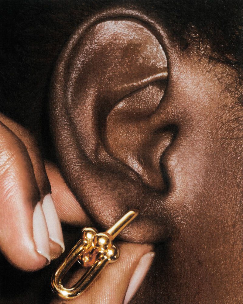 Close-up shot of a dark-skinned woman's ear with a gold earring featuring a unique 'U' shaped design being held by manicured fingers.