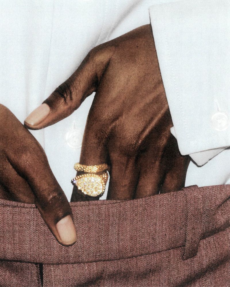 Close-up of a hand with brown skin, adorned with a large gold ring, resting in the front pocket of mauve trousers.