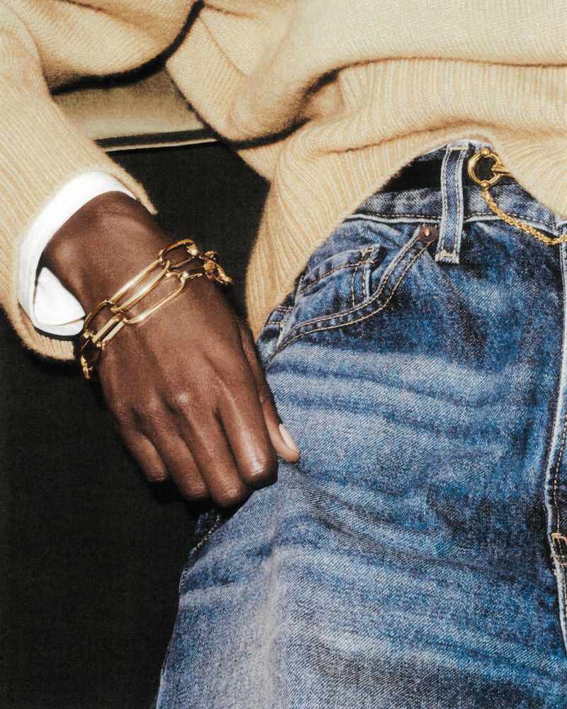 Close-up studio shot of a dark-skinned arm wearing a chunky gold chain bracelet, paired with a beige sweater and faded blue jeans.