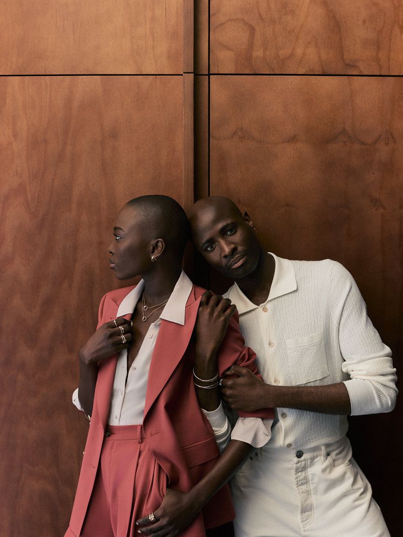 Two young Black adults pose indoors against a wood panel wall; the female wears a rust-colored suit and the male wears white.