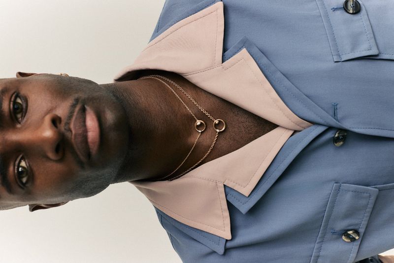 Close-up of a dark-skinned adult male wearing a blue shirt and gold necklaces against a white background in a studio setting.
