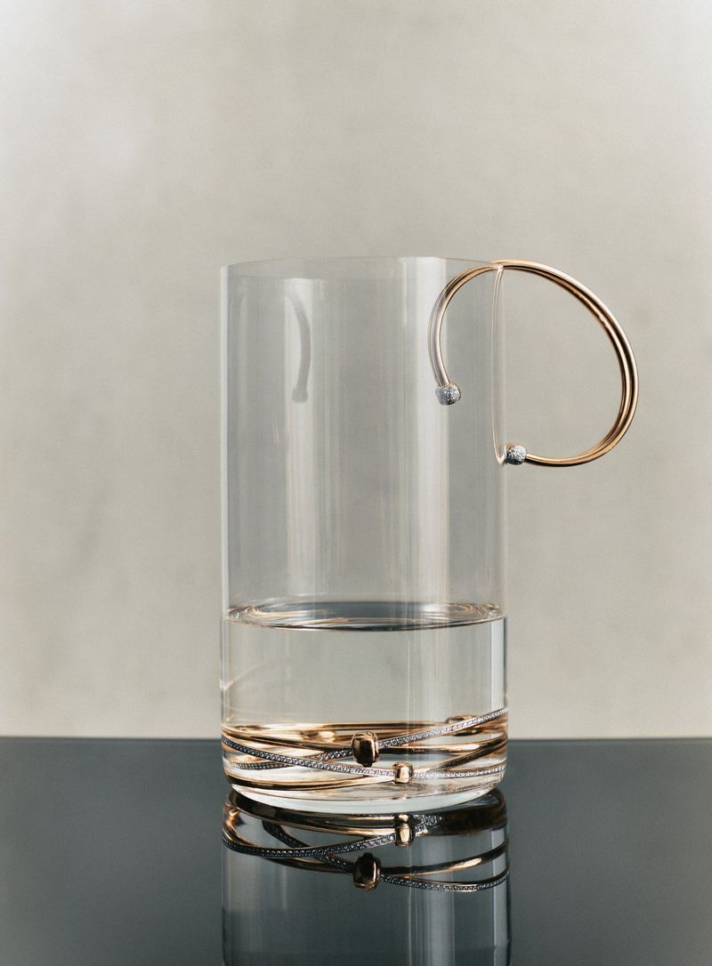 Glass mug filled with water with a gold handle sits on a reflective black surface against a cream-colored wall.