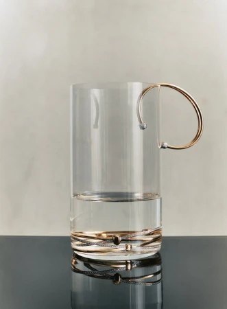 Glass mug filled with water with a gold handle sits on a reflective black surface against a cream-colored wall.