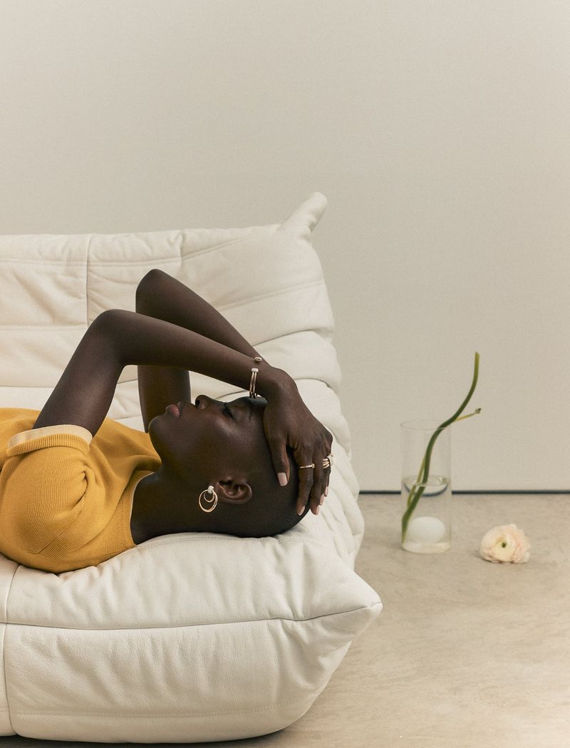 An adult Black woman lying on a white cushioned sofa, wearing a yellow shirt and silver jewelry, with a minimalist background and floral accents.