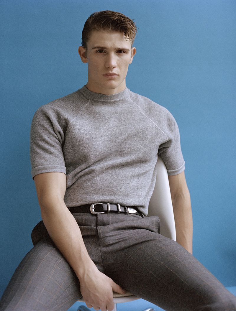Man with fair skin wearing gray sweater and pants posing while sitting on a chair against blue background in a medium eye-level shot.