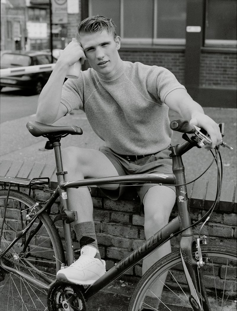 Black and white portrait of a blonde adult male on a bicycle in front of a brick wall outdoors in daylight with neutral expression.