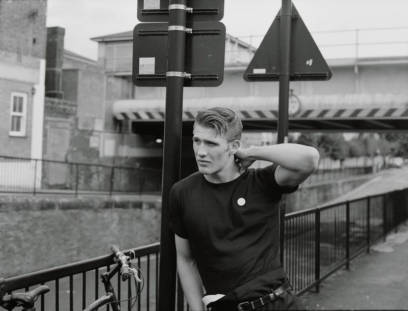 Monochrome portrait of a stylish young adult man posing with a bike in an urban setting with buildings and a bridge in the background.