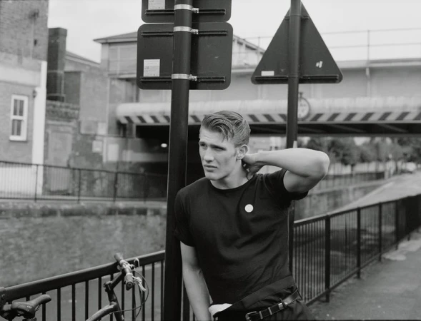 Monochrome portrait of a stylish young adult man posing with a bike in an urban setting with buildings and a bridge in the background.