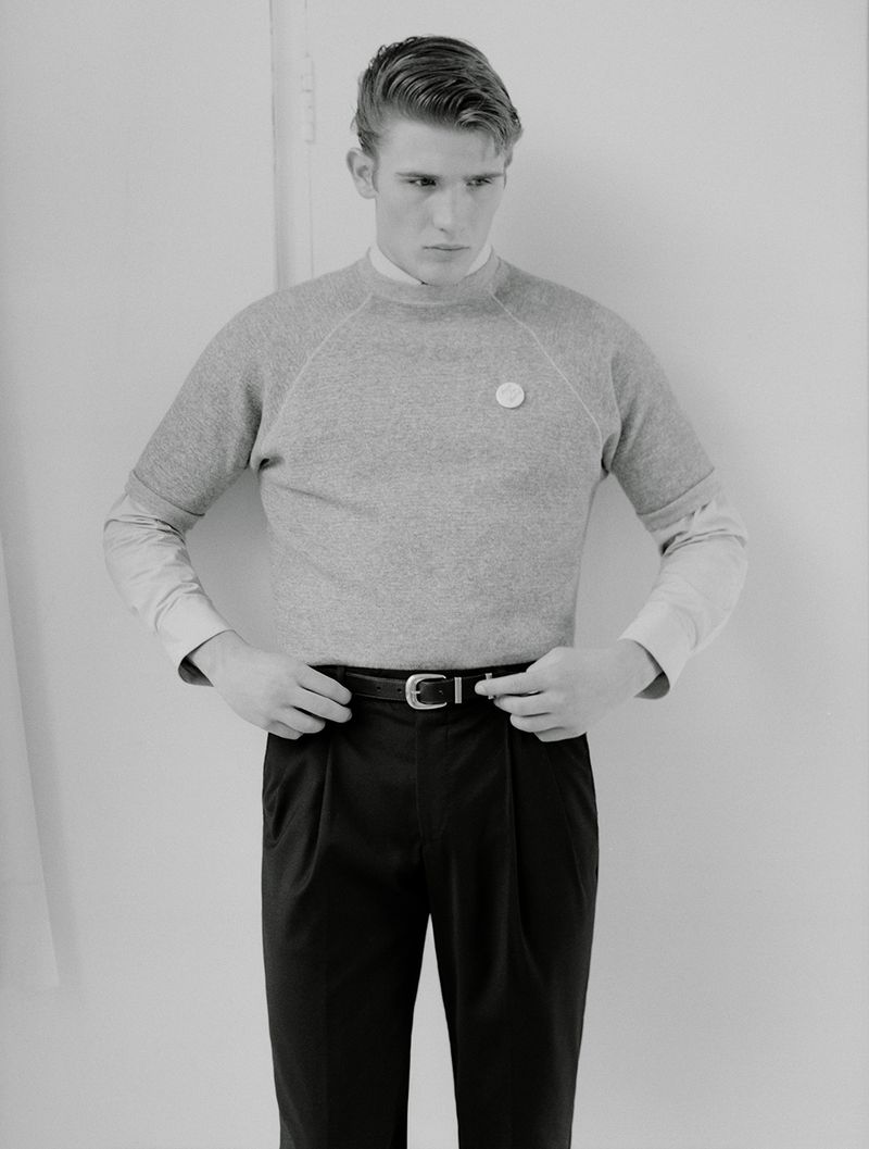 Monochrome portrait of a young adult male in a casual grey sweater and dark trousers, standing against a plain white backdrop.