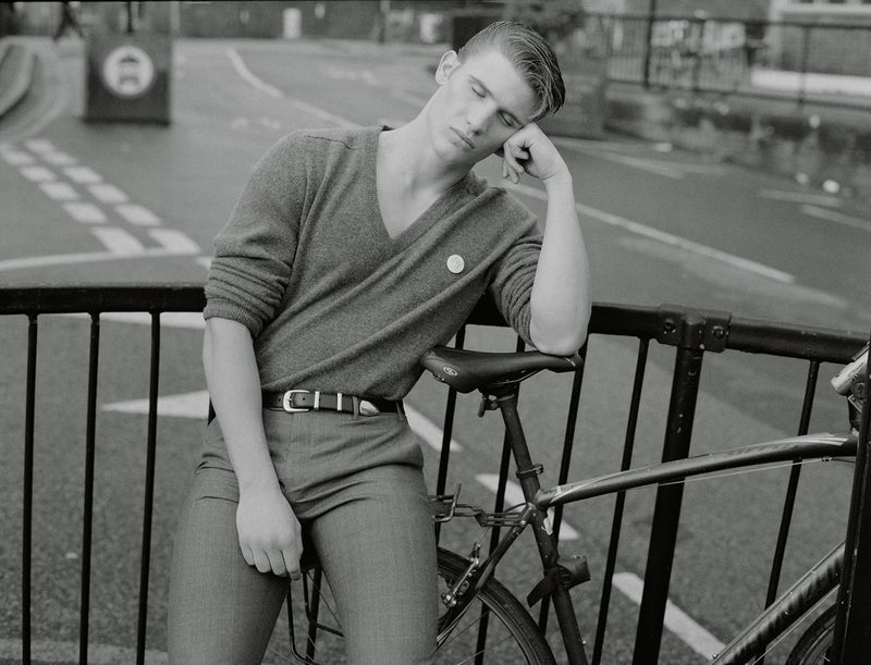 Black and white shot of a young man in sweater and slacks leaning on a bicycle on an urban bridge, eyes closed.