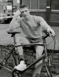 A young male model holding a bottle while seated on a bicycle outdoors against a brick wall background