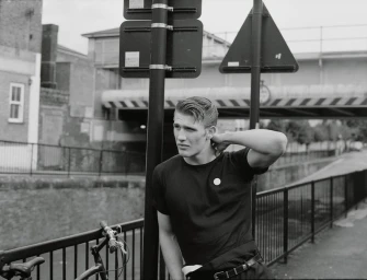 Black and white portrait of a man in a dark t-shirt leaning against a street sign pole near a bridge.