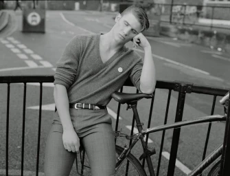 Monochrome portrait of a male model leaning against a bicycle behind a metal railing on an empty street.
