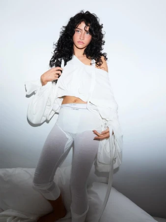 A young woman with curly hair poses indoors wearing white cotton crop top and legging set with visible logo waistband in a bright studio.